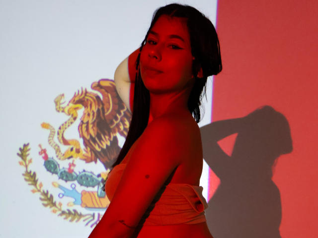 Photograph of a young woman with wet hair, ZoeWinfrey, standing in red light, shadowed against a Mexican flag backdrop. She's topless, small breasts visible, looking at camera with sultry expression.