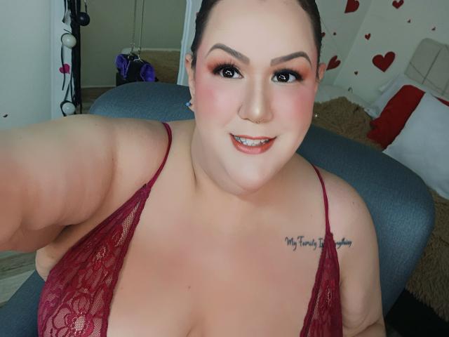 Fotografía de una GiiannaMorrys regordeta, con piel pálida, cabello oscuro y lápiz labial rojo, sonriendo a la cámara. Ella lleva una blusa roja de encaje que deja ver parte de su escote, y tiene un tatuaje en el hombro derecho. Increíblemente atractivo/a..