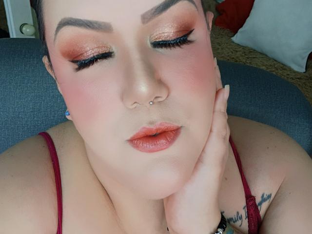 Nahaufnahme von GiiannaMorrys mit intensivem, schimmerndem orangefarbenem Lidschatten, rosa Rouge und rotem Lippenstift. Sie legt ihre Wange auf ihre Hand, wobei ein Nasenpiercing und ein rotes Tanktop zu sehen sind. Ihre Augen sind halb geschlossen, und sie strahlen eine entspannte, verführerische Ausstrahlung aus..