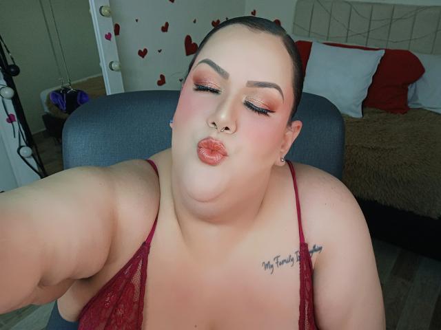 Close-up selfie of GiiannaMorrys with her eyes closed, puckering her lips for a kiss. She's wearing a red lace top, showcasing her plump, fair-skinned body with a tattoo on her shoulder.