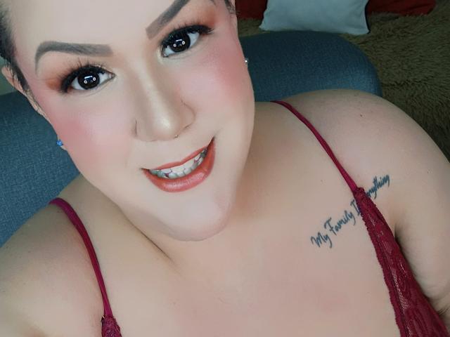 Close-up photo of GiiannaMorrys, a plus-sized woman with fair skin, dark eyebrows, and red lipstick. She's smiling, showing teeth, with a red spaghetti strap top and a tattoo on her right shoulder. Fucking sexy.