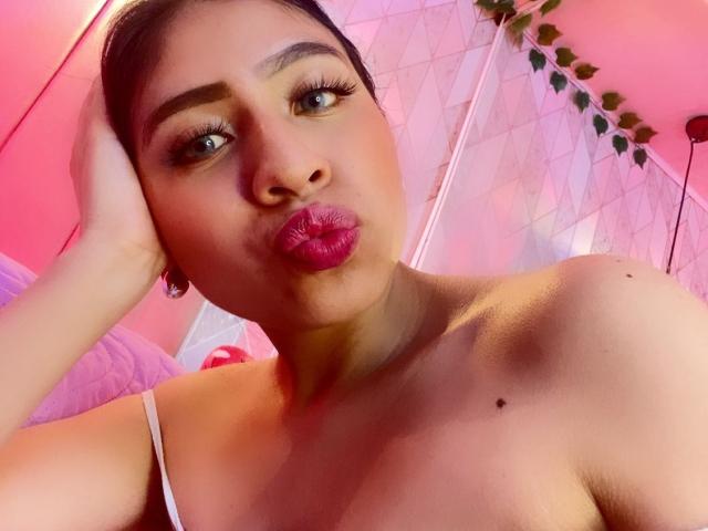 Close-up of ZoeRedds, brown-skinned woman with dark hair, pouty pink lips, and brown eyes. She's making a kissing face, head tilted, hand on cheek. Pink and white background. Sexual tension.