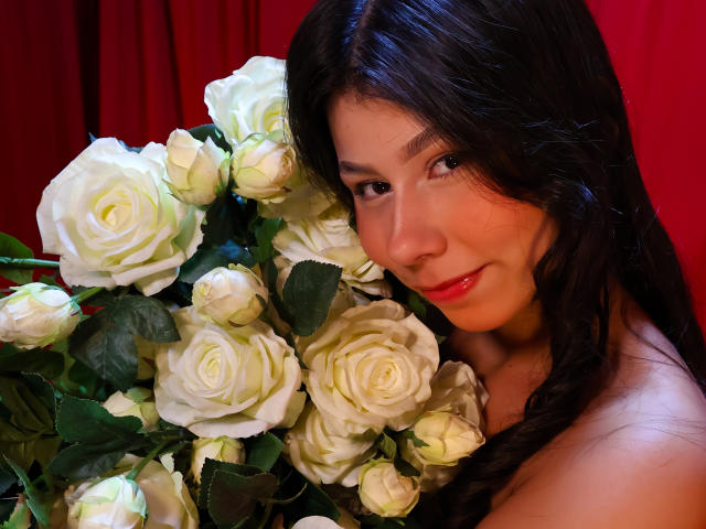 Fotografie eines Nahporträts von ZoeWinfrey mit langen, schwarzen Haaren, die an einem großen Strauß weißer Rosen lehnt. Roter Hintergrund. ZoeWinfrey hat ein leichtes Lächeln, rote Lippen. Verdammt heiß..