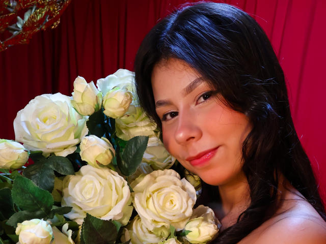 Fotografie einer attraktiven asiatischen Frau mit langen, schwarzen Haaren, die an einem Strauß weißer Rosen lehnt. Ihr Gesicht ist gerötet, mit einem vielversprechenden Lächeln. Helle, rote Hintergrundfarbe. Helle Beleuchtung.