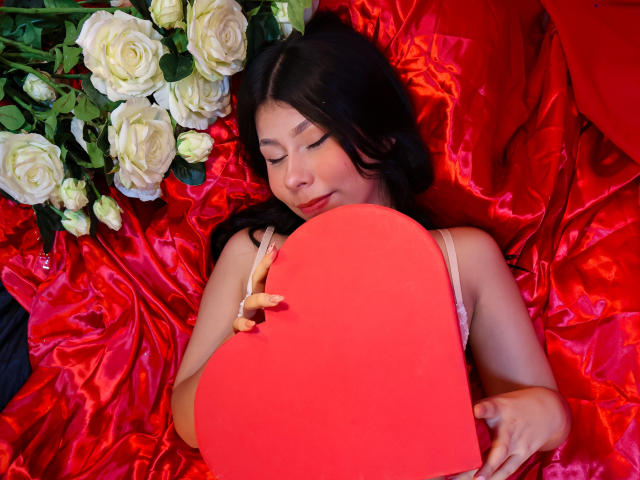 Zoe Winfrey sdraiata su lenzuola di seta rossa, che tiene in mano un grande cuore rosso, con gli occhi chiusi, circondata da rose bianche. I suoi capelli scuri le ricadono sulle spalle. Un'atmosfera incredibilmente romantica..