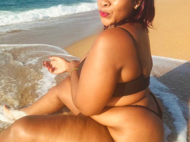 QueenBebe mit dunkelbrauner Haut, rotem Lippenstift und einem schwarzen Bikini sitzt auf einem sandigen Strand mit Wellen, mit dem Rücken zum Betrachter, und blickt Über die Schulter geschaut. Das Sonnenlicht betont ihre Kurven und erzeugt eine sinnliche, verführerische Atmosphäre..