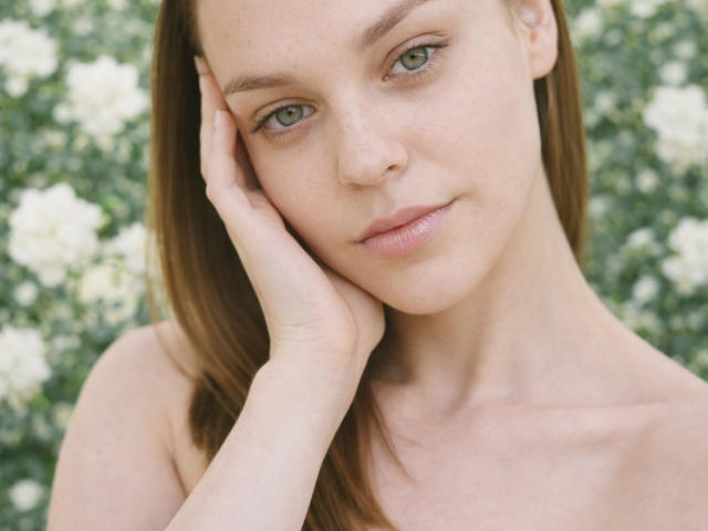 Nahaufnahme von LolaaQueenn, hellhäutig mit grünen Augen, braunen Haaren und rosafarbenen Lippen, die sich mit ihren Fingern das Gesicht berührt. Rechte Hand. Der Hintergrund ist unscharf und zeigt grüne und weiße Blumen. Absolut wunderschön..