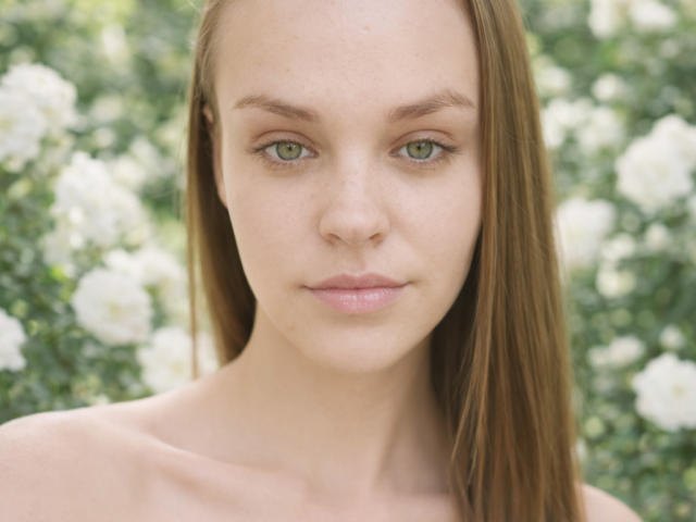 Fotografia em close-up de LolaaQueenn, de pele clara, com olhos verdes, cabelo castanho liso, nua, olhando para a câmera, luz suave. Fundo verde e branco desfocado, tensão sexual sutil, expressão neutra, luz natural..