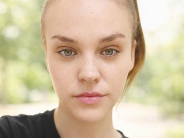 Close-up of LolaaQueenn with fair skin, green eyes, and light brown hair pulled back. She has a neutral expression, glossy pink lips, and is wearing a black top. Blurred green background.