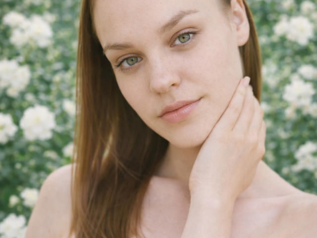Close-up of LolaaQueenn with pale skin, green eyes, and brown hair, touching her neck with her right hand. She has a subtle smirk, wearing nothing, with a blurred white flower background.
