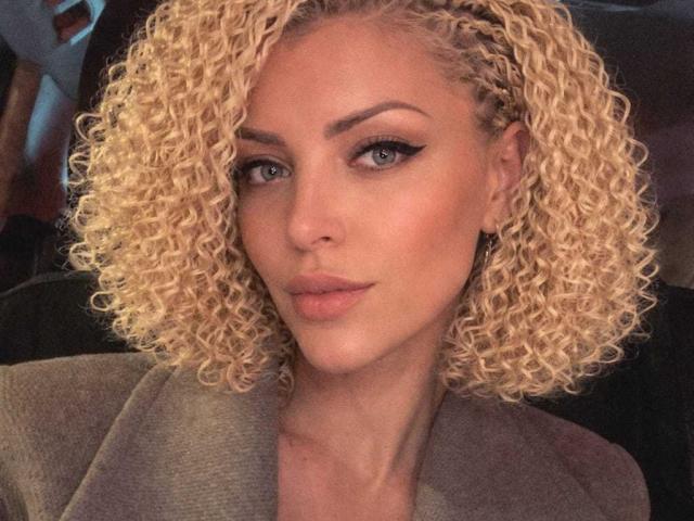 Primo piano di Axinia con ricci biondi corti e ben definiti, pelle chiara e un trucco occhi nero molto marcato. Ha labbra piene leggermente dischiuse e indossa un blazer grigio. Incredibilmente sexy, sguardo intenso..