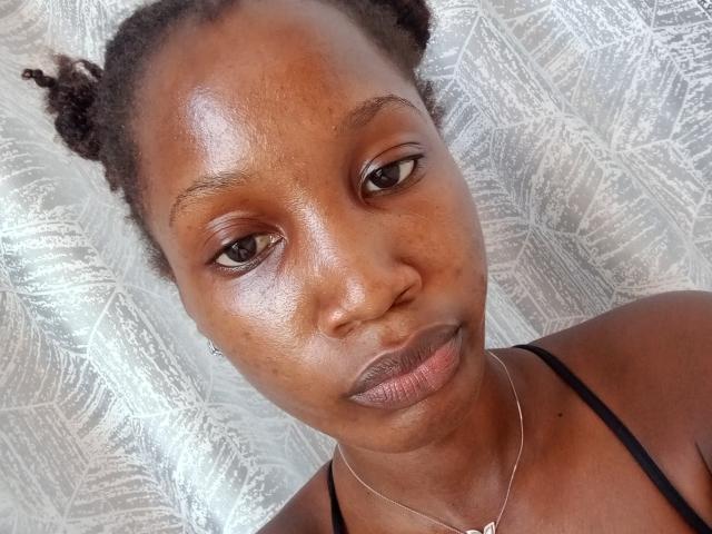 Close-up of SexxyRedd with dark brown skin, shiny face, and black hair in two small braids. She wears a thin black strap top and a delicate silver necklace. Fucking gorgeous.