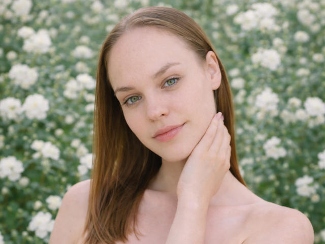 Foto van LolaaQueenn, met een lichte huid, recht bruin haar, blauwe ogen en een subtiele glimlach. Ze raakt haar nek aan met haar linkerhand, op de achtergrond zijn wazige witte bloemen te zien. Heel aantrekkelijk..