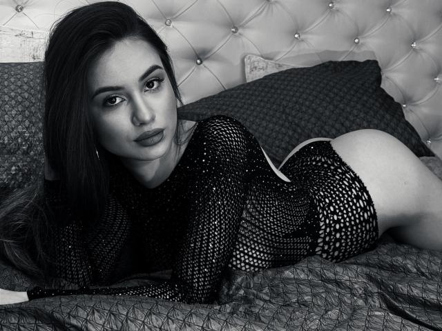 AmiellaRose, allongée sur un lit, portant un haut en maille noire qui expose ses fesses, regarde directement l'appareil photo avec sensualité. yeux, cheveux foncés, tête de lit capitonnée, photo en noir et blanc, sexy, provocateur.