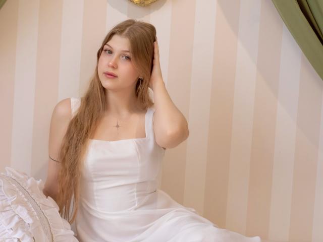 StellaLane está sentada em uma cama, vestindo um vestido branco com decote quadrado, cabelos castanhos longos e um colar discreto. A mão dela toca a cabeça, e os olhos azuis claros se desviam. Fundo suave com listras..