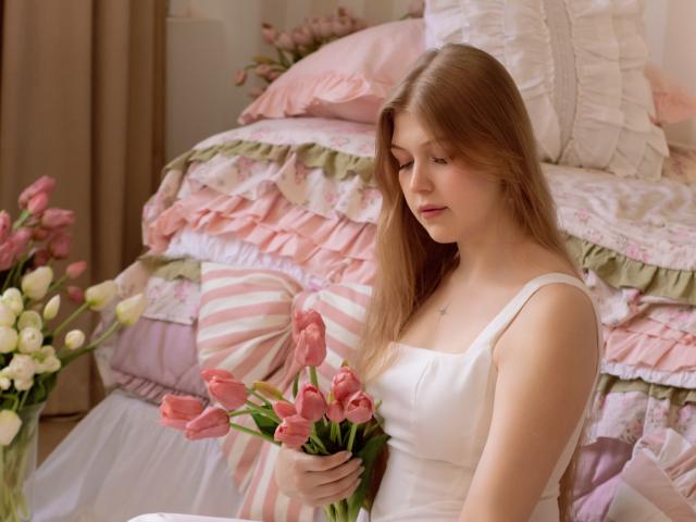 StellaLane, avec ses longs cheveux bruns, est assise sur un lit rose et blanc à volants, tenant des tulipes roses. Elle porte une robe blanche, le regard baissé, et une douce lumière naturelle projette des ombres..