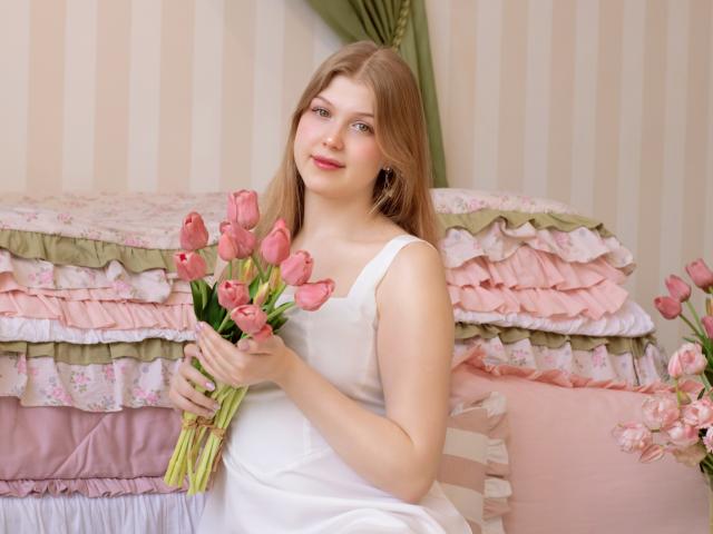 StellaLane met lang bruin haar, die roze tulpen vasthoudt, gekleed in een witte jurk, zittend op een roze en witte bed met ruches, zacht licht, bloemenachtergrond, verleidelijke blik, licht roze wangen..