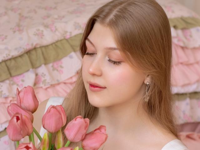 Close-up of StellaLane with straight brown hair, holding pink tulips, eyes closed, wearing a white top, pink floral pillows in the background, soft pink lipstick, earring on right ear.