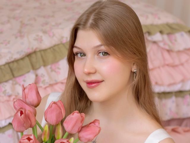 StellaLane, een vrouw met een lichte huid en lang bruin haar, houdt roze tulpen vast, draagt een witte top, en ligt op een bed met roze bloemenprints op het beddengoed. in de achtergrond. Zachte glimlach, subtiele seksuele spanning, lichte make-up, oorbellen.