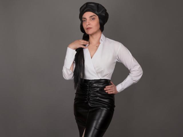 Xrystal, com cabelos longos e pretos presos em um coque, vestindo uma camisa branca de botões e calças de couro pretas justas. Mão na cintura, outra mão tocando o pescoço. Fundo cinza. Pose confiante e sensual..