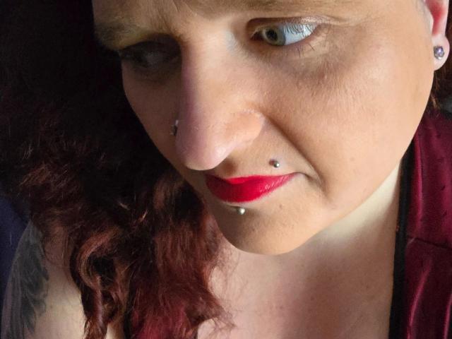 Primo piano di hotsunny20-sex, una donna bianca con rossetto rosso, piercing al naso e alle labbra, capelli ricci castano chiaro, sguardo intenso. sfondo scuro, luce intensa sul viso, leggero sorriso, espressione seducente.