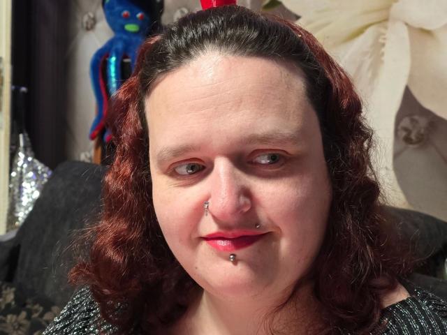 Primer plano de hotsunny20-sex, una mujer de piel clara con cabello rizado rojo, perforación en la nariz y perforación en el labio, vestida con algo brillante. superficie negra, con una estatua azul y tela blanca en el fondo.