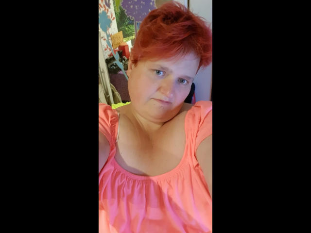 Hotsunny20-sex, eine rothaarige Frau mit kurzen Haaren, die ein pinkes Oberteil trägt, lehnt sich zurück und lächelt leicht, wobei sie dekolleté, leuchtende Farben, unscharfer Hintergrund mit bunten Objekten, lässige Pose.