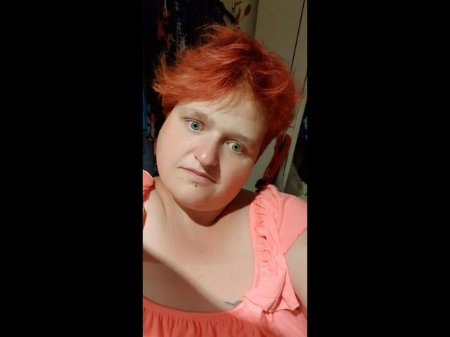 Photograph of hotsunny20-sex with short, messy red hair, fair skin, and blue eyes. Wearing a pink, ruffled top. Sunlight casts shadows on her face, creating a sensual, intense look.