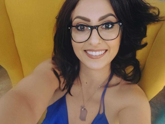 Close-up photo of LisaCroftt, smiling with white teeth, wearing black-framed glasses, blue halter top, and a purple crystal necklace, seated against a yellow chair, showing a fucking hot look.