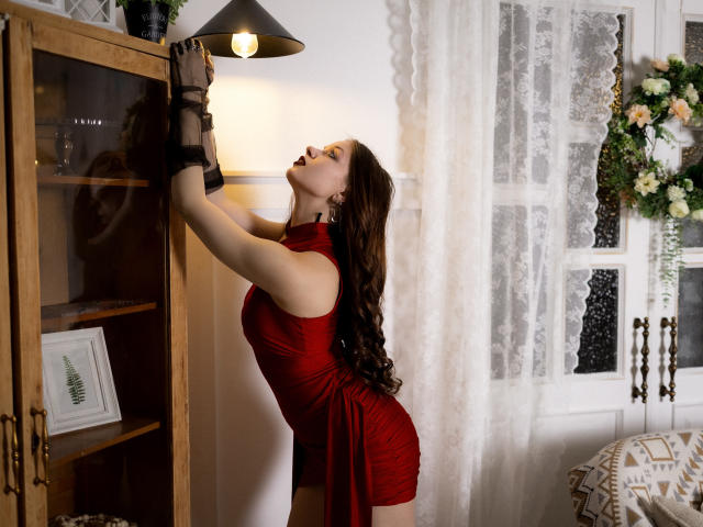 BethCutie, eine kurvige Frau mit langen, dunklen Haaren, trägt ein eng anliegendes rotes Kleid, schwarze Handschuhe und beugt sich vor, um eine Lampe zu justieren. auf einem Holzkabinett. Verdammt sexy. Blumenarrangement auf der rechten Seite..