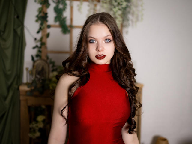 BethCutie, com olhos azuis marcantes, vestindo um vestido vermelho justo sem mangas, com longos cabelos negros ondulados e batom vermelho escuro. O fundo apresenta uma planta verde e uma prateleira de madeira. Incrivelmente atraente..