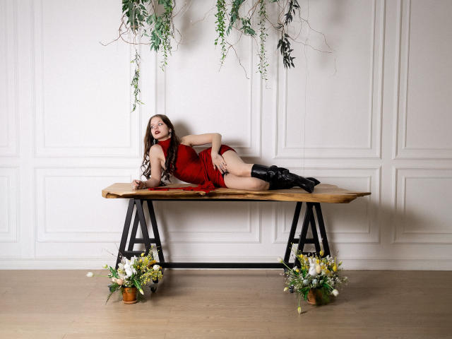 BethCutie lies on a wooden bench with black metal legs, wearing a red dress, legs spread, black panties down, floral plants around, white paneled wall, seductive pose, sexual tension.