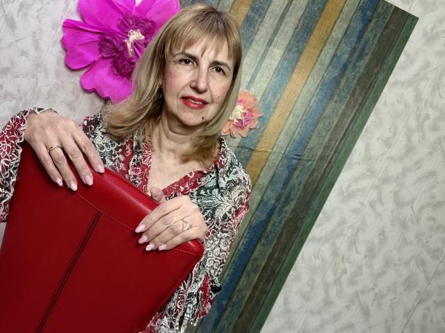 PattySanny, eine Frau mittleren Alters mit blonden Haaren und rotem Lippenstift, hält ein rotes Kissen. Sie trägt ein Oberteil mit einem schwarz-weißen Muster und rosa Blumen im Haar. Verdammt heiß..