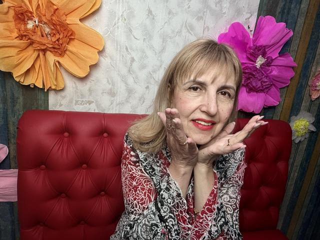 PattySanny sits on a red tufted couch, hands framing her face with a flirtatious smile. She's wearing a patterned shirt, flanked by large orange and pink flowers. Fucking sexy.