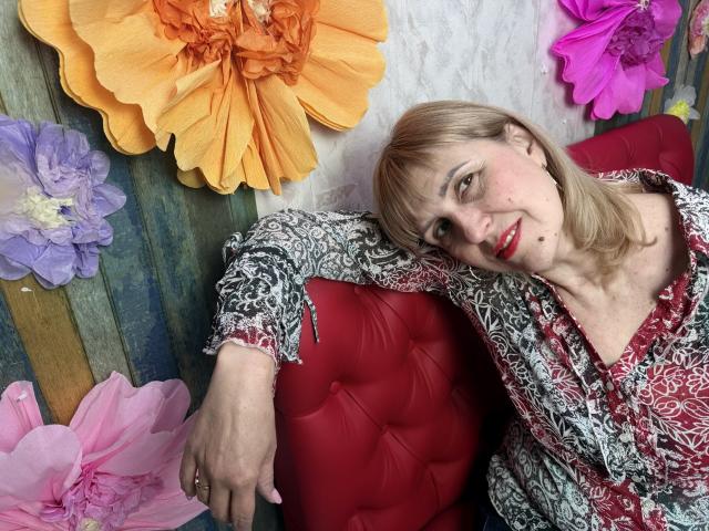 PattySanny è sdraiata su un divano rosso con decorazioni in tessuto, circondata da grandi fiori di carta colorati. Ha il rossetto rosso, i capelli biondi e una maglietta a righe rosse e bianche. Completamente rilassato, guarda la telecamera..