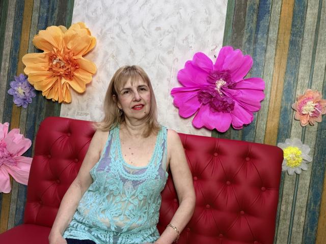 PattySanny, une femme d'âge moyen avec des cheveux blonds mi-longs, est assise sur un canapé rouge capitonné, vêtue d'une robe bleu clair en dentelle. en haut.

de grandes fleurs en papier, aux couleurs vives d'orange, de rose et de violet, l'entourent..