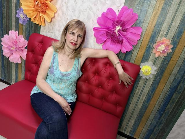 PattySanny sitzt auf einem roten, gepolsterten Sofa, hinter ihr befinden sich große, bunte Papierblumen. Sie trägt ein blaues Spitzen-Tanktop und schwarze Jeans, und ihr Arm ruht auf der Rückenlehne. Verdammt heiß..