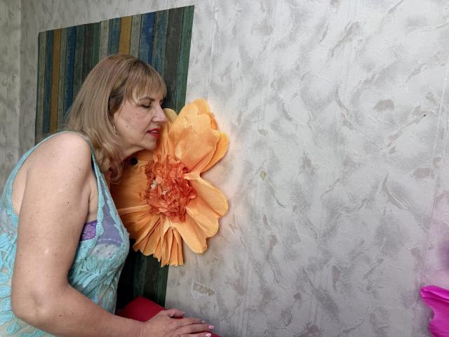 PattySanny, une femme d'âge moyen aux cheveux bruns clairs, est appuyée contre un mur blanc texturé, le regard fixé sur un grand objet orange. fleur. Elle porte un haut bleu à motifs, et on peut voir ses ongles roses..