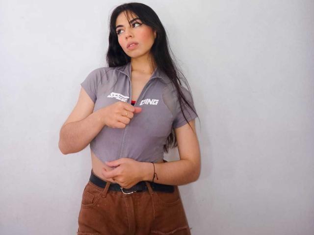 Katherin Johns è in piedi di fronte a un muro bianco, guardando di lato con un'espressione leggermente seducente. Indossa una maglietta grigia aderente a maniche corte con la scritta "PANGA" e dei pantaloni marroni. La sua mano è sul petto, le dita stringono leggermente la maglietta. Capelli lunghi e neri, pelle abbronzata, corporatura esile. I suoi occhi sono socchiusi e le labbra leggermente dischiuse, emanando una sottile tensione sessuale..