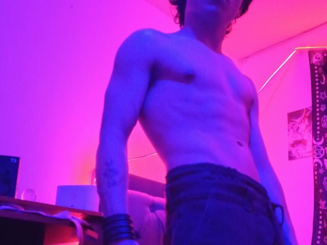 Shirtless HiddenX with a lean, muscular build, small nipples, and a tattoo on the left arm, wearing dark pants, standing in purple-pink lighting, with wristbands on the right wrist.
