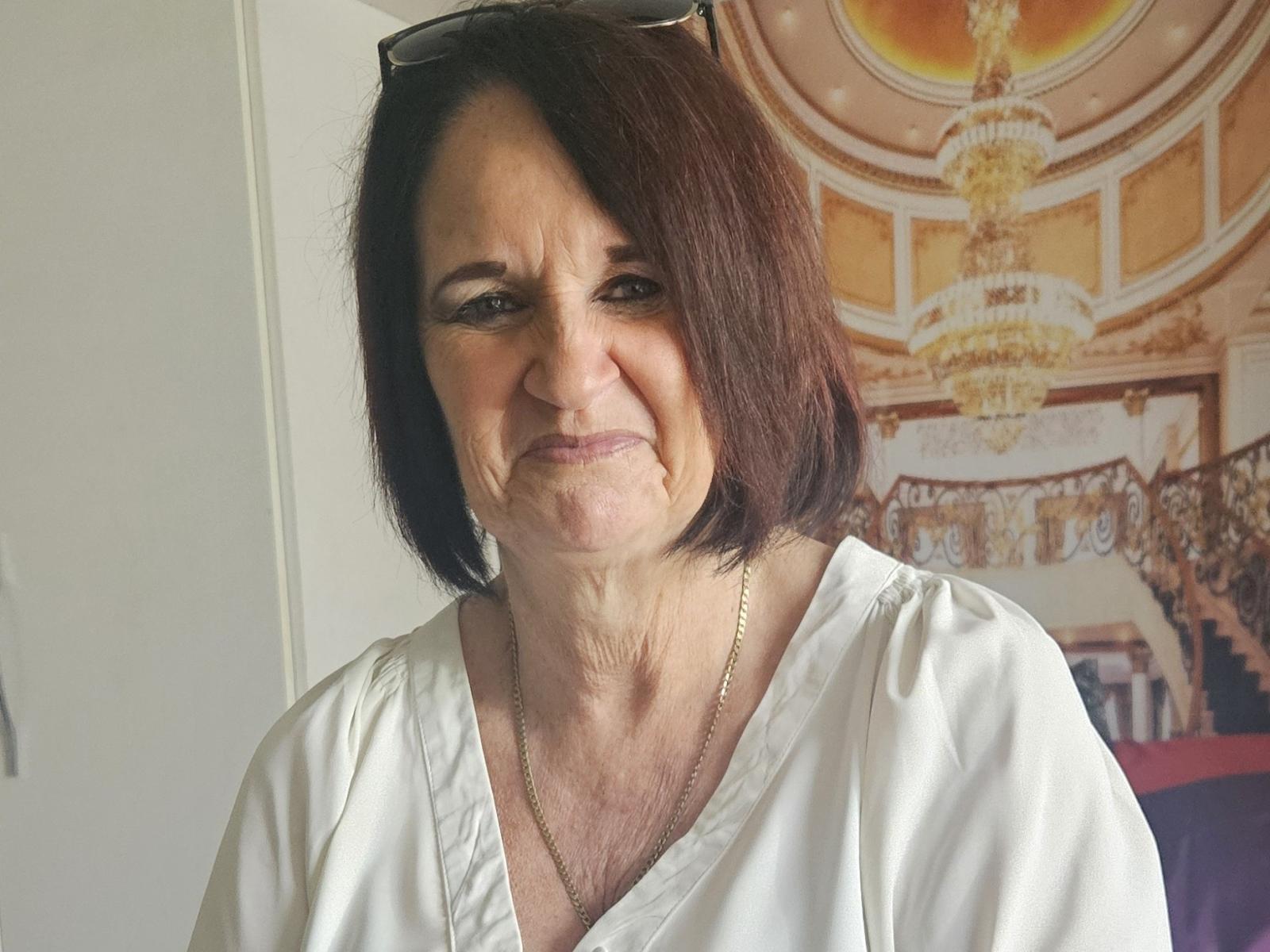 LuciousGrace, mittleren Alters, mit schulterlangem, schwarzem Haar, trägt eine weiße Bluse mit V-Ausschnitt und steht vor einer verzierten wandgemälde mit einem Chandelier. Sie hat einen ernsten Gesichtsausdruck und eine goldene Halskette. Eine richtig ernste Ausstrahlung..