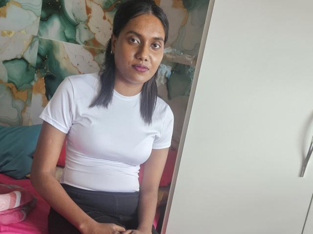 IndianSkies with dark hair, white t-shirt, and black pants, sitting, looking at camera, small boobs, slight smile, pink bed, white door, green and white marble wall.