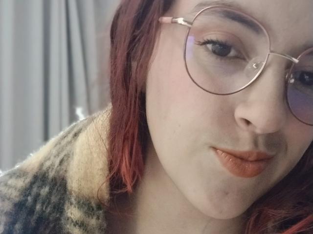 Close-up photo of LaiaJonnes with fair skin and red hair, wearing round glasses and a fur-trimmed coat. She's looking at the camera with a subtle smirk, lips slightly parted.