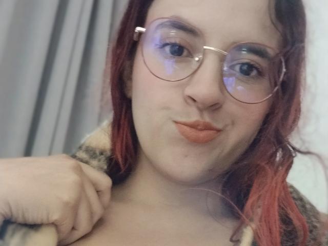 Close-up of LaiaJonnes, fair-skinned with red hair, wearing round glasses and orange lipstick, looking at the camera with a slight smile. Her left hand grips a fabric near her shoulder.