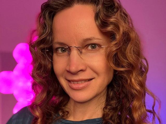 Close-up of AnnikaRose-hot, a woman with curly brown hair, wearing glasses, and a dark blue shirt. Pink neon lights in the background. Her expression is confident, slightly seductive.