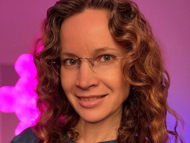 Close-up of AnnikaRose-hot, smiling with curly brown hair, wearing glasses, against a pink neon light background. Her expression exudes subtle sexual tension, enhanced by the soft, warm lighting.