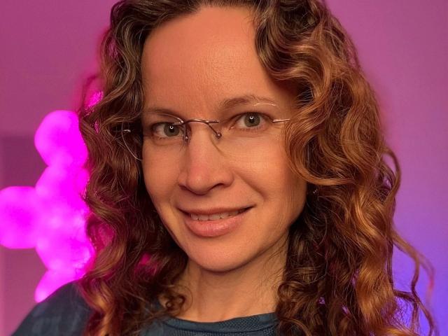 Close-up photograph of AnnikaRose-hot, a woman with curly brown hair, wearing glasses, and a dark blue shirt. Pink neon light in the background, smiling, soft facial features, slight blush.