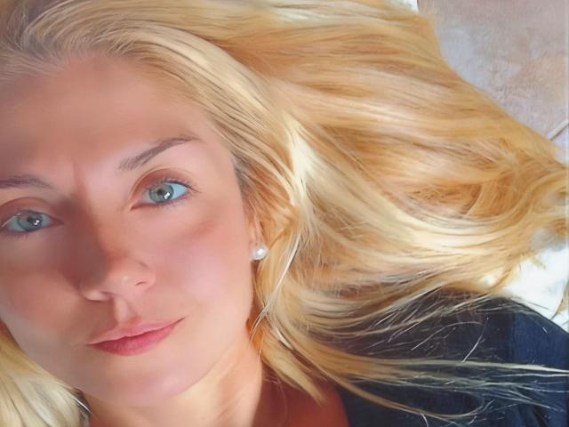 Fotografía de primer plano de MiaAsh, con cabello rubio extendido, ojos azules, piel clara, un sutil labial rojo, ropa negra y blanca. Fondo. Mirada intensa y atractiva, ligera sonrisa, pose relajada, luz natural..