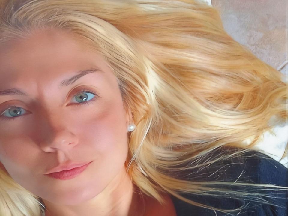 Fotografía de primer plano de MiaAsh, con cabello rubio extendido, ojos azules, piel clara, un sutil labial rojo, ropa negra y blanca. Fondo. Mirada intensa y atractiva, ligera sonrisa, pose relajada, luz natural..