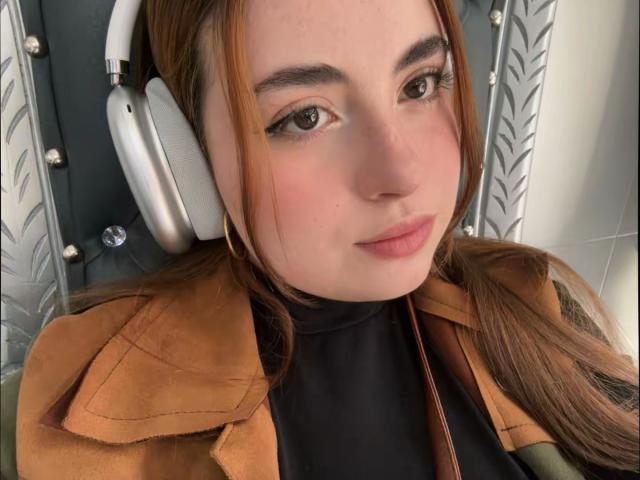 Fotografía de primer plano de VicluX, una mujer de piel clara y cabello castaño, que lleva unos auriculares blancos grandes, un abrigo marrón y pantalones negros. Con un cuello alto, mirando directamente a la cámara con una sonrisa sutil. Tensión sexual evidente..